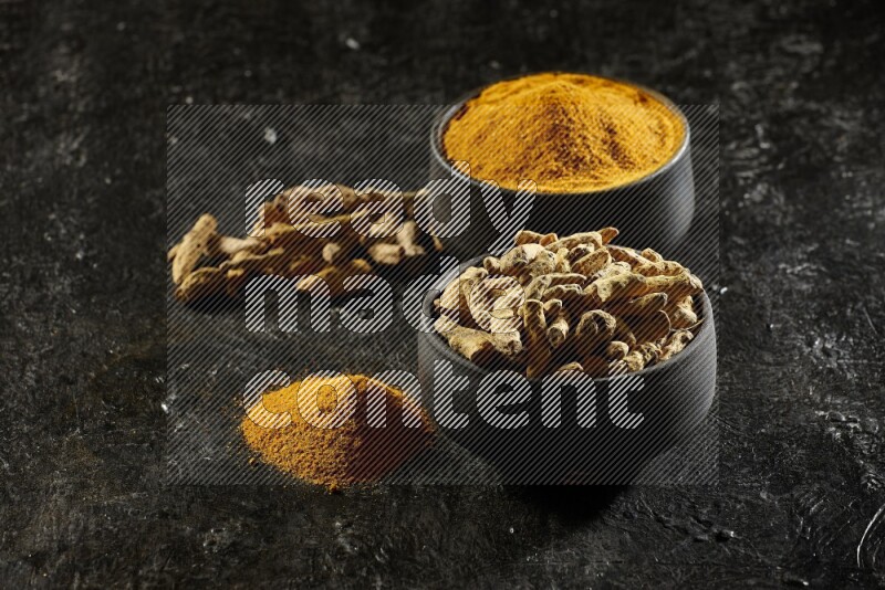 2 pottery black bowls, one full of turmeric powder and the other full of dried turmeric whole fingers on a textured black flooring