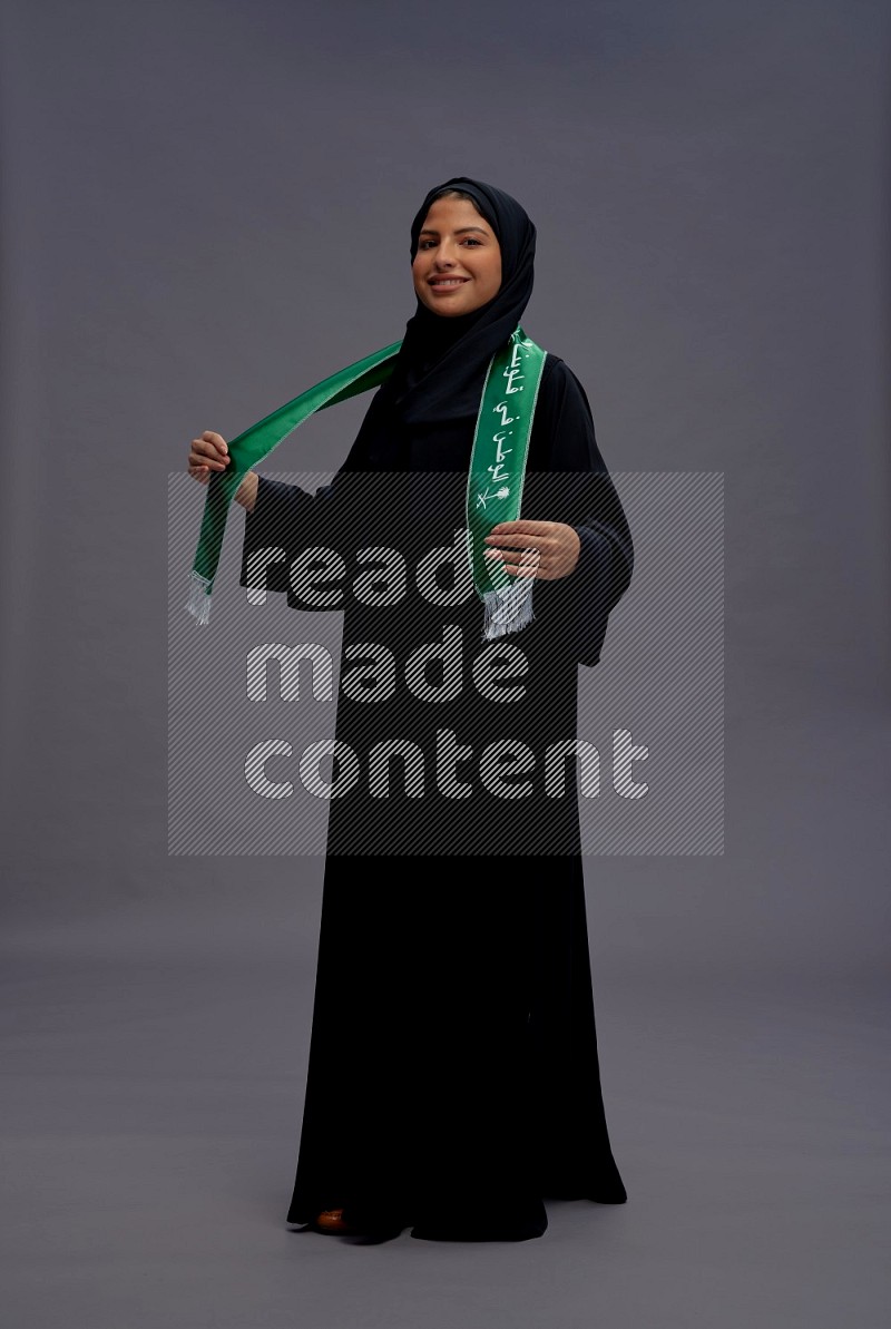 Saudi woman wearing Abaya standing holding Saudi flag on gray background