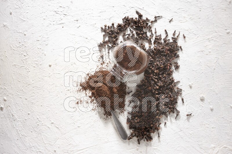 A flipped glass spice jar and a metal spoon full of cloves powder and powder came out of the jar with cloves spread on textured white flooring