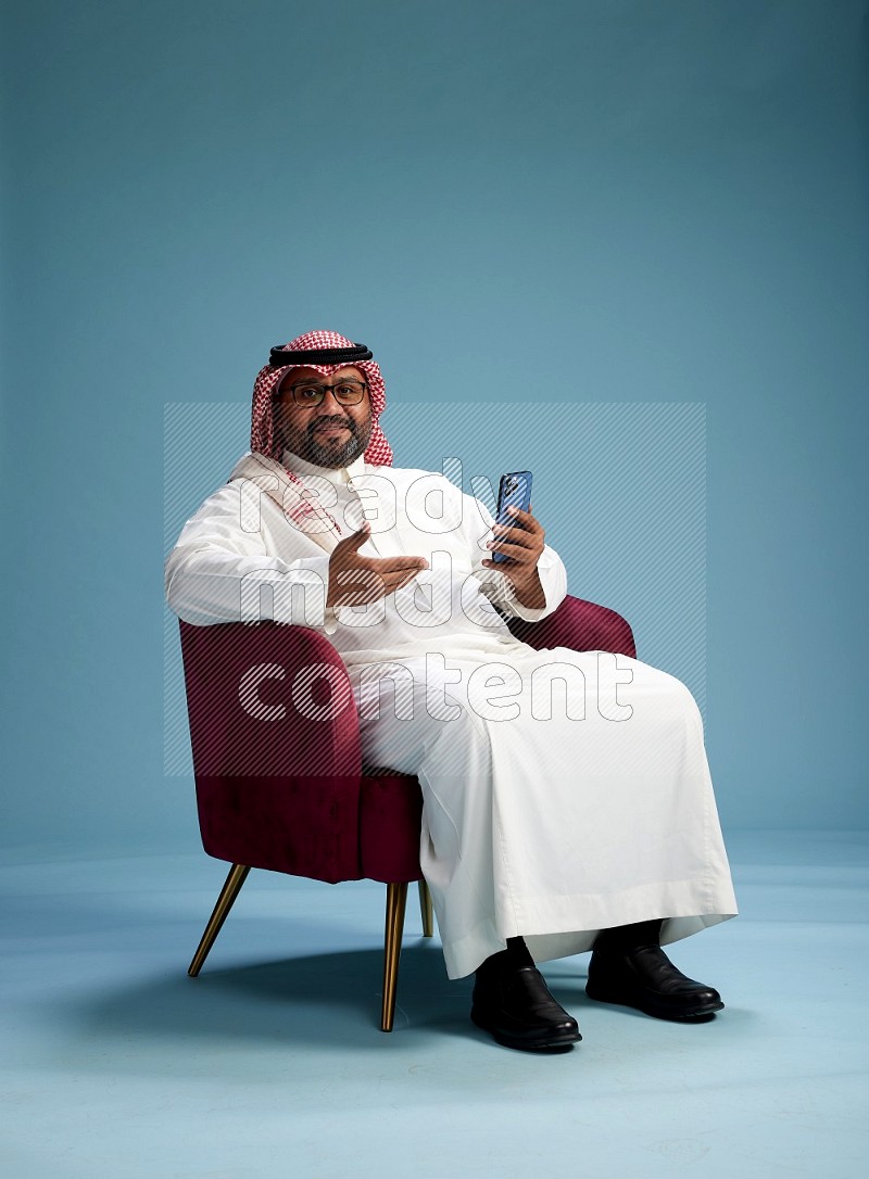 Saudi Man with shimag sitting on chair texting on phone on blue background