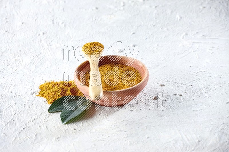 A wooden bowl and wooden spoon full of turmeric powder on textured white flooring