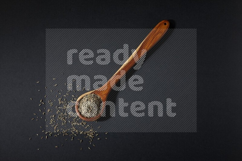 A wooden ladle full of cumin seeds on black flooring