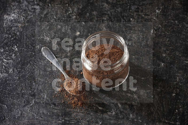 وعاء زجاجي ممتلئ ببودرة القرنفل مع ملعقة معدنية على أرضية سوداء