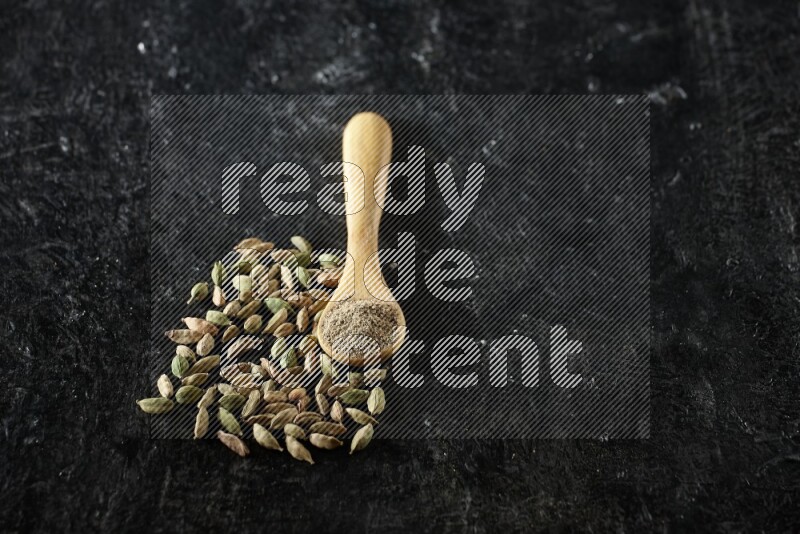 A wooden spoon full of cardamom powder and cardamom seeds next of it on textured black flooring