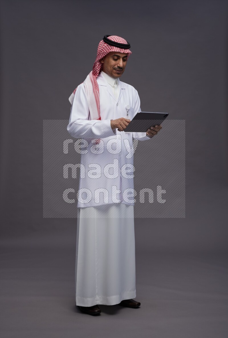 Saudi man wearing thob with lab coat and shomag with pocket employee badge standing working on tablet on gray background