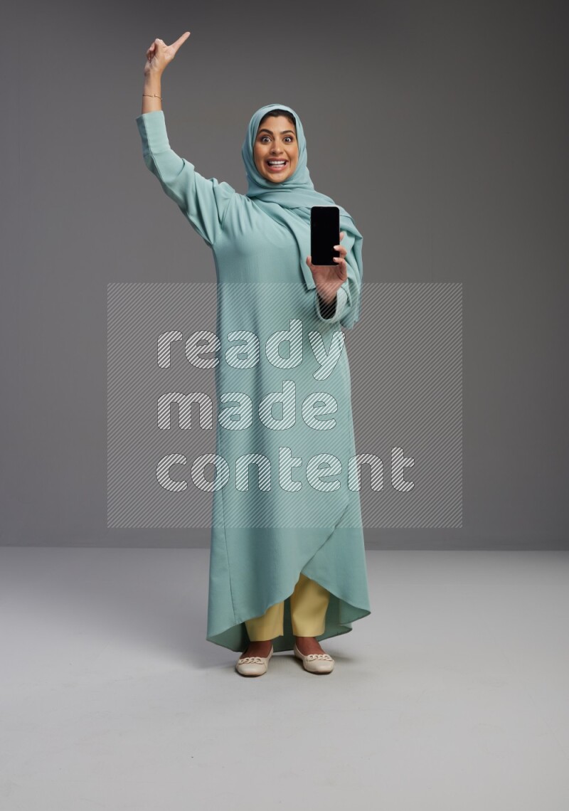 Saudi Woman wearing Abaya standing showing phone to camera on Gray background