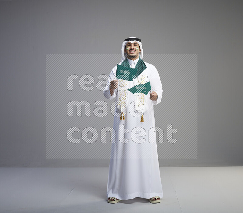 A saudi man standing wearing thob and white shomag with face painting and saudi flag scarf and holding small saudi flag on gray background
