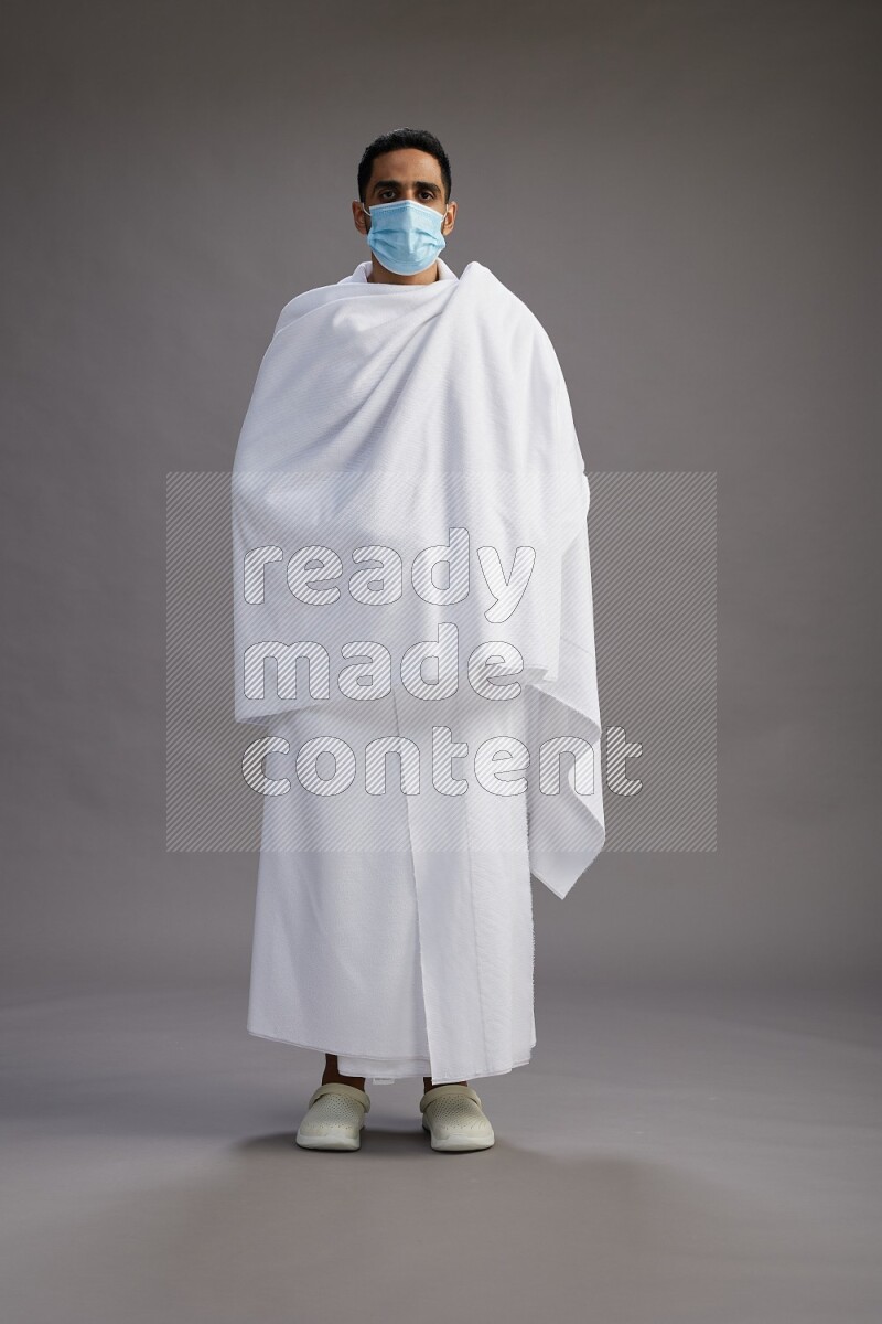 A man wearing Ehram with face mask Standing Interacting with the camera on gray background