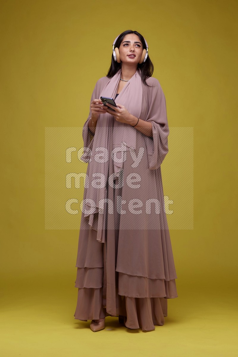 A Saudi woman Listing to Music on a Yellow Background wearing Brown Abaya