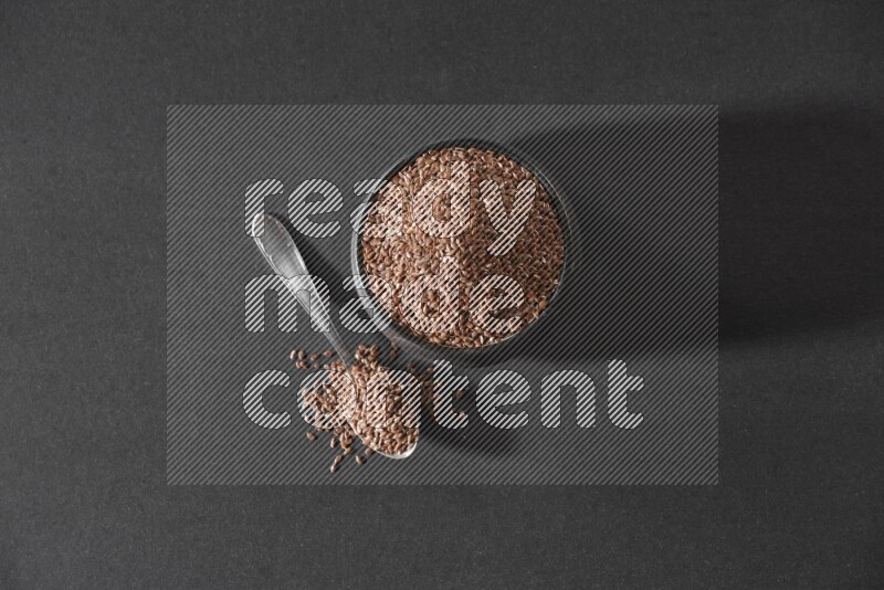 وعاء فخاري أسود ممتلئ بحبوب بذر الكتان مع ملعقة معدنية ممتلئة على أرضية سوداء