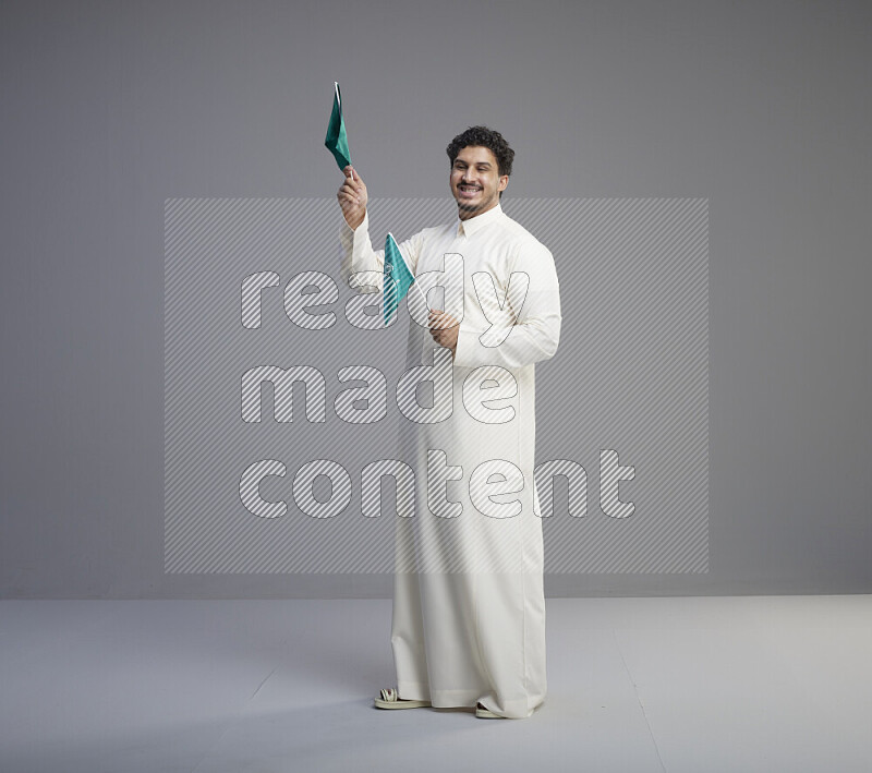 A Saudi man standing wearing thob holding small Saudi flag on gray background