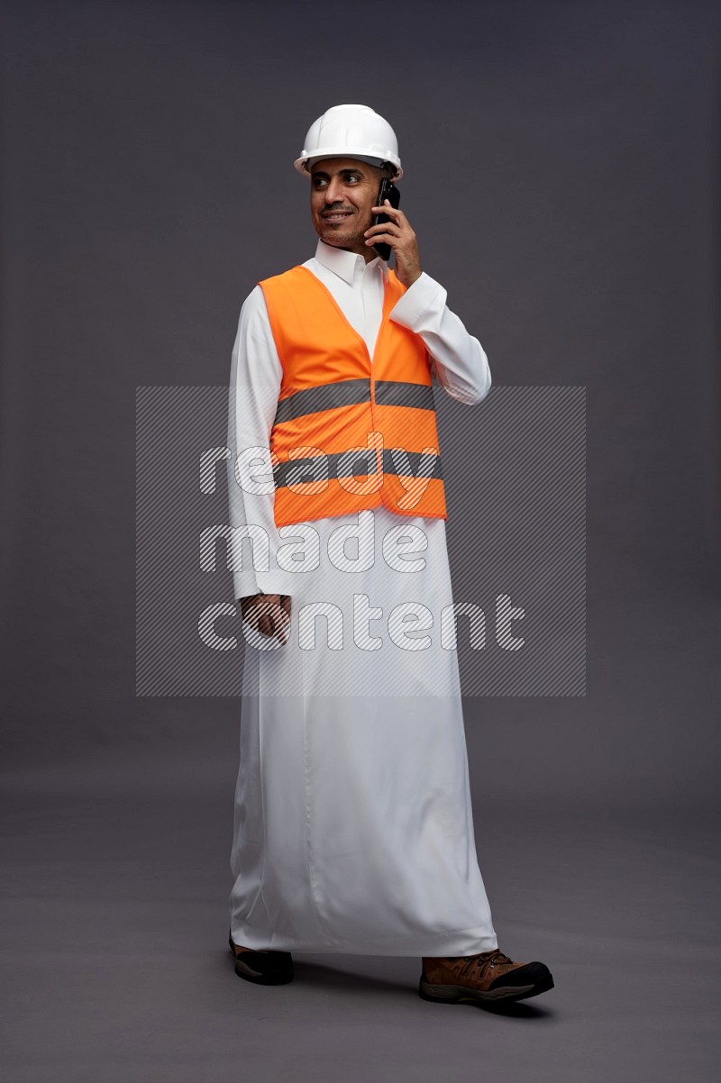 Saudi man wearing thob with engineer vest standing talking on phone on gray background