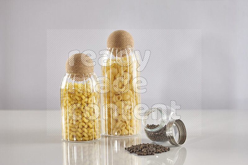 Raw pasta in glass jars with black peppers on light grey background