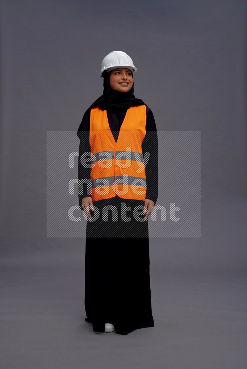 Saudi woman wearing Abaya with engineer vest standing interacting with the camera on gray background