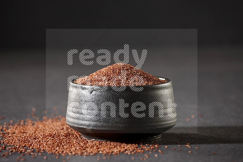 A black pottery bowl full of garden cress seeds surrounded by seeds on a black flooring