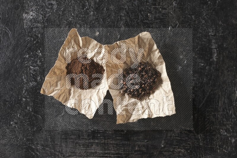 Cloves powder and cloves in 2 crumpled pieces of paper on a textured black flooring