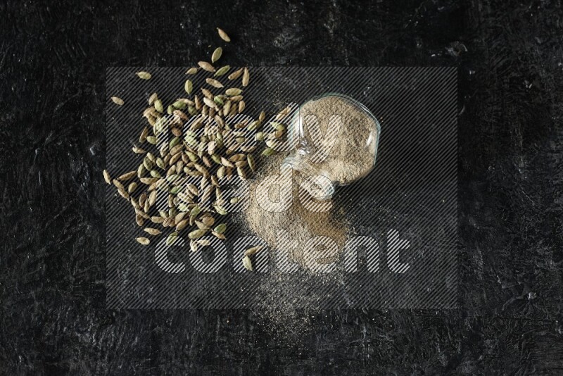 A flipped glass spice jar full of cardamom powder and powder spilled out of it with cardamom seeds spreaded on textured black flooring