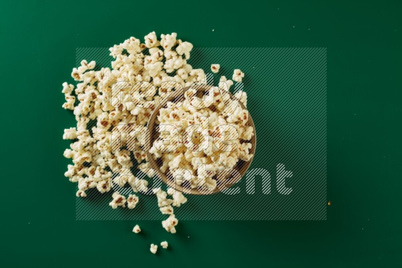 An off white ceramic bowl full of popcorn with popcorn beside it on a green background in a top view shot