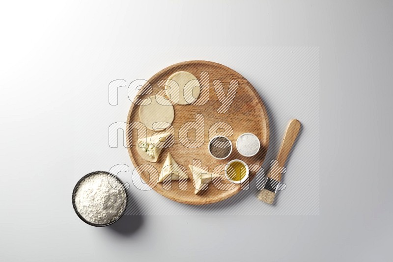 two closed sambosas and one open sambosa filled with cheese while flour, salt, black pepper and oil with oil brush aside in a wooden dish on a white background