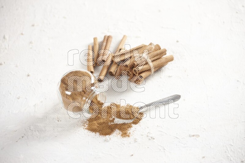 Flipped herbs glass jar full of cinnamon powder with a metal spoon full of powder and cinnamon sticks on a textured white background