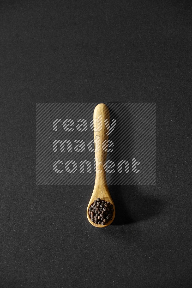 A wooden spoon full of black pepper on a black flooring