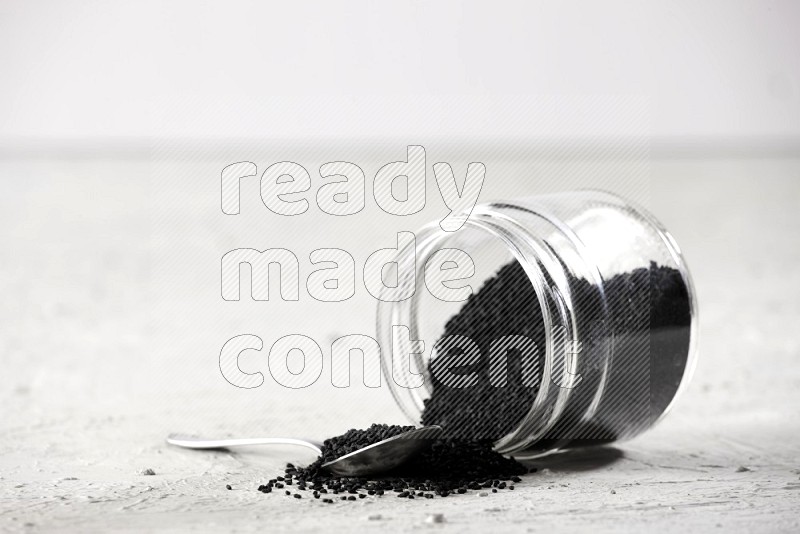 A glass jar and a metal spoon full of black seeds and the jar flipped and seeds spread on a textured white flooring