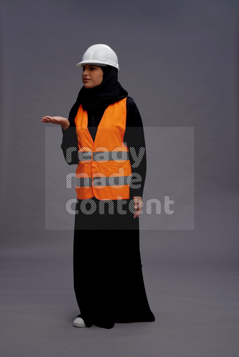 Saudi woman wearing Abaya with engineer vest standing interacting with the camera on gray background