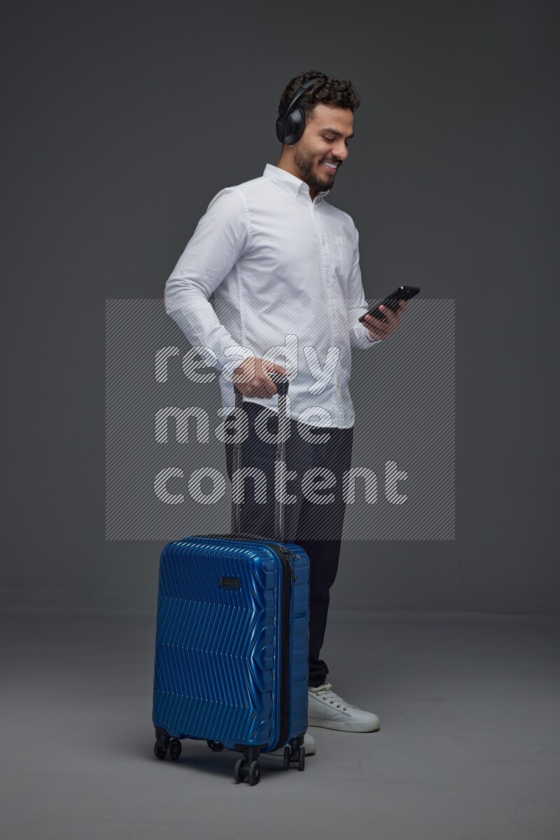 A man wearing smart casual and headphone holding luggage eye level on a gray background