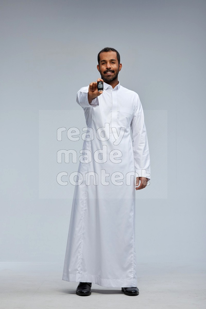 Saudi man wearing thob and shomag standing holding car key on gray background