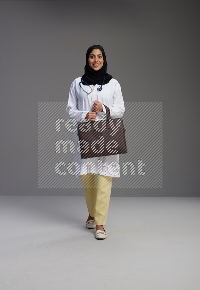 Saudi woman wearing lab coat with stethoscope standing holding bag on Gray background