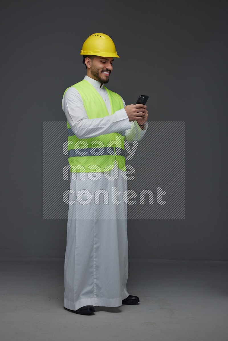 A Saudi man wearing Thobe with a yellow safety vest and white helmet standing and using his phone eye level on a gray background