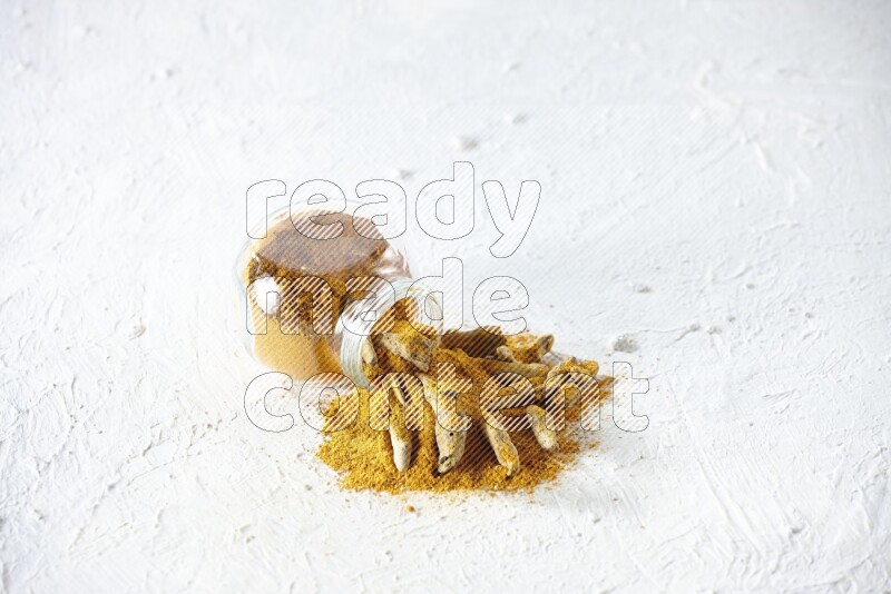 A flipped glass spice jar full of turmeric powder and powder spilled out of it with dried whole fingers on textured white flooring