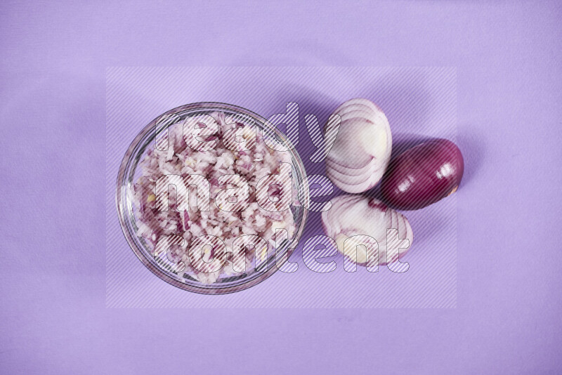 A glass bowl full of chopped red onions with halved onions beside it on purple background