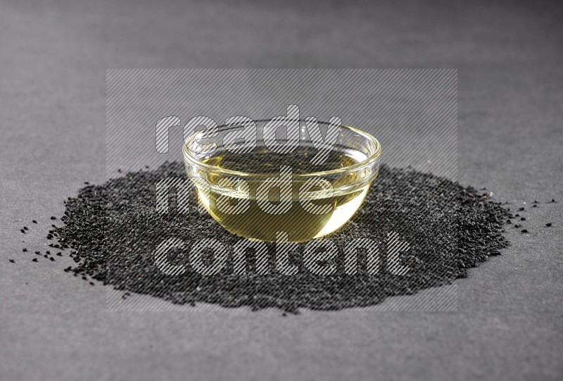 A glass bowl full of black seeds oil surrounded by the seeds on a black flooring