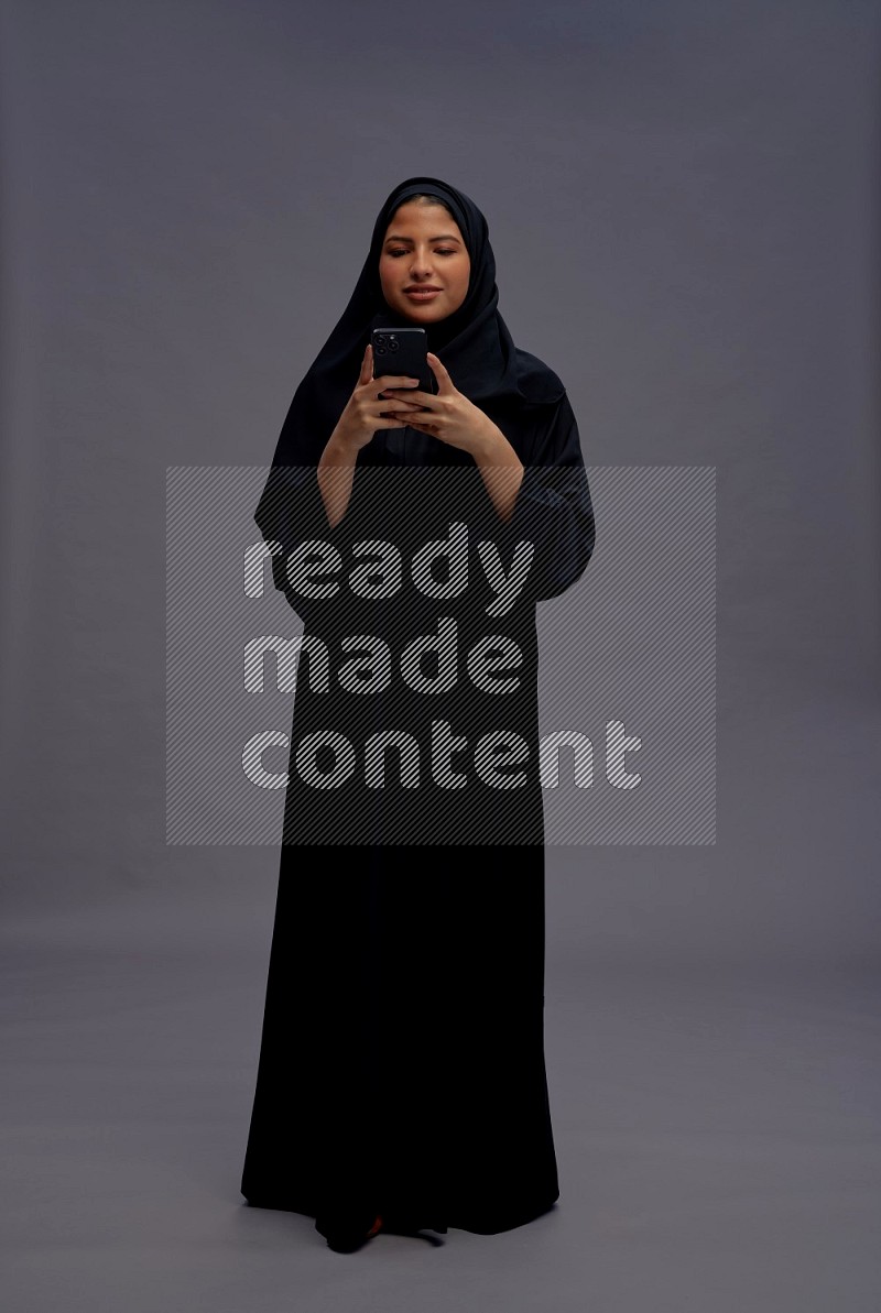 Saudi woman wearing Abaya standing texting on phone on gray background