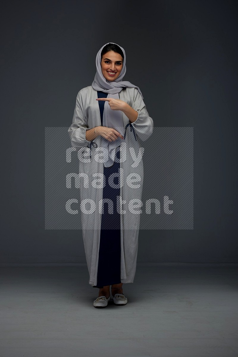 A Saudi woman wearing a light gray Abaya and head scarf standing and pointing in different directions eye level on a grey background