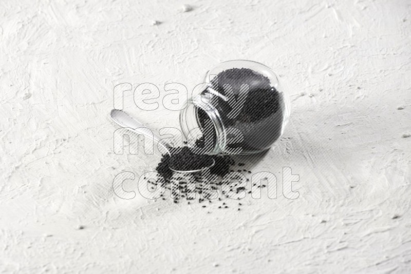 A glass spice jar and a metal spoon full of black seeds and jar is flipped with fallen seeds on a textured white flooring in different angles