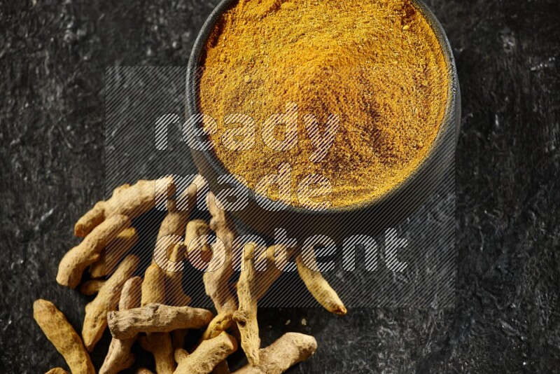 A black pottery bowl full of turmeric powder and dried turmeric whole fingers next of it on textured black flooring
