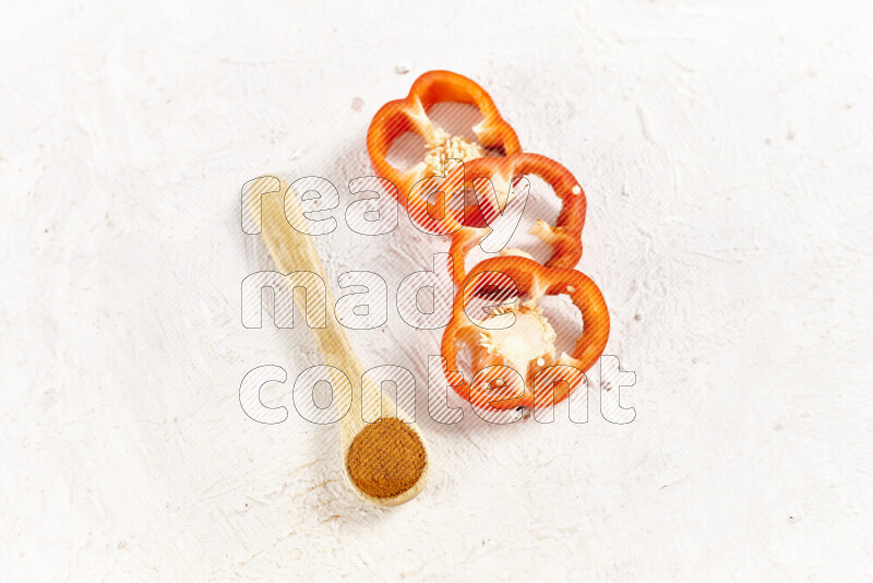 A wooden spoon full of ground paprika powder with red bell pepper slices beside it on white background