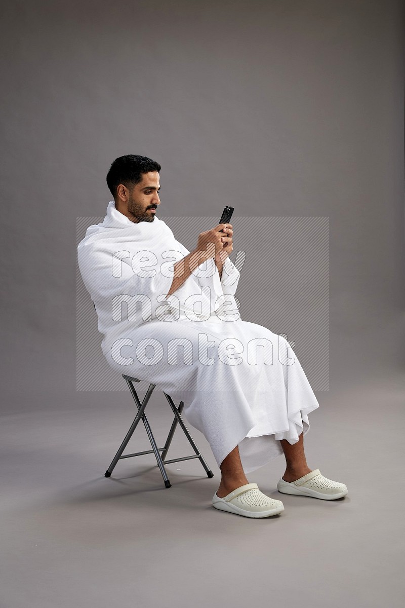 A man wearing Ehram sitting on chair texting on phone on gray background
