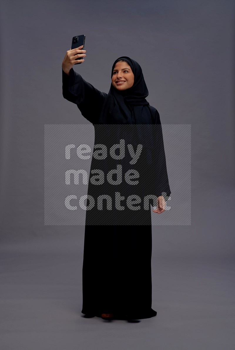 Saudi woman wearing Abaya standing taking selfie on gray background