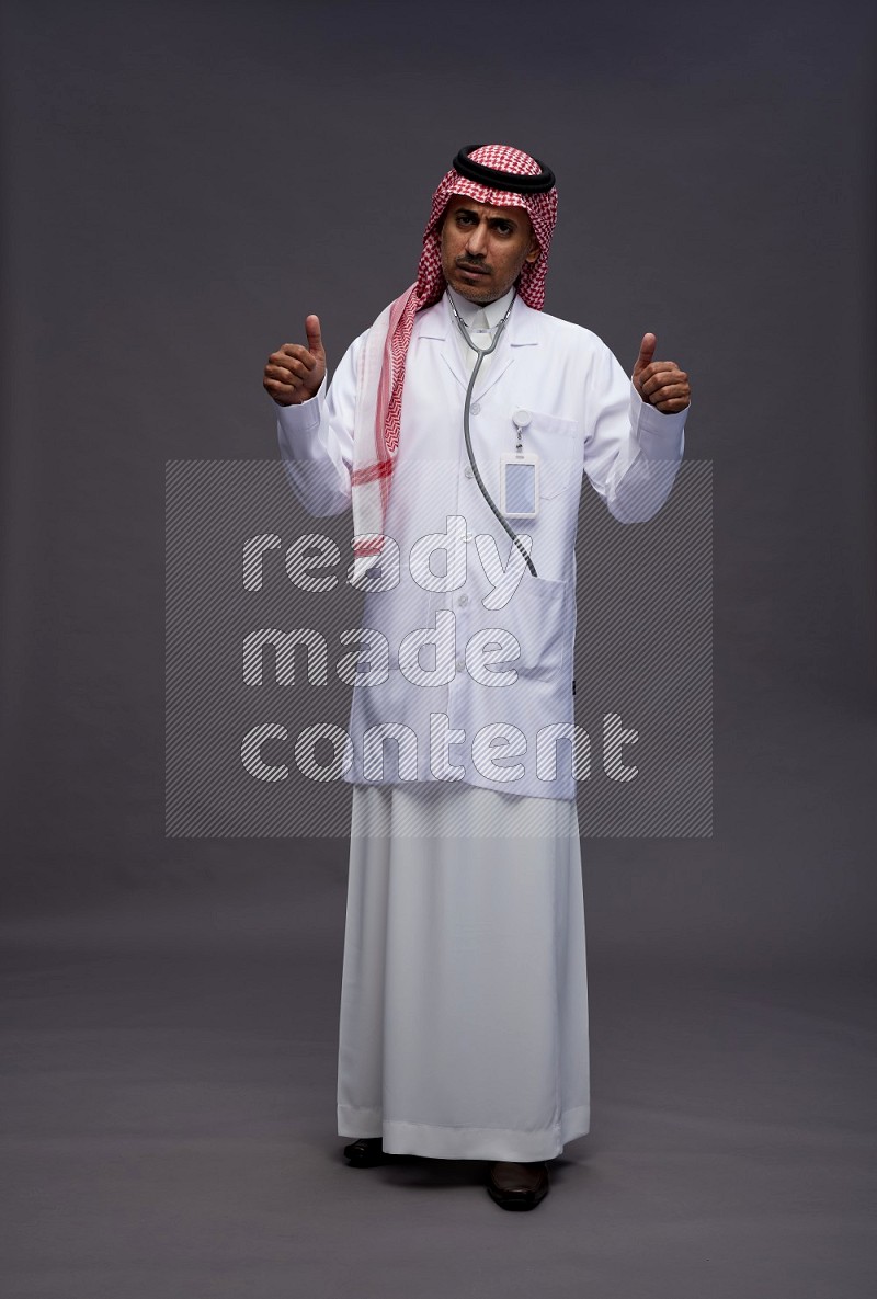 Saudi man wearing thob with lab coat and shomag with pocket employee badge with stethoscope standing interacting with the camera on gray background