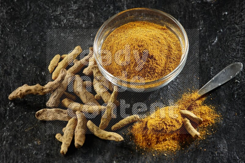 A glass bowl and a metal spoon full of turmeric powder and dried whole fingers next of them on textured black flooring