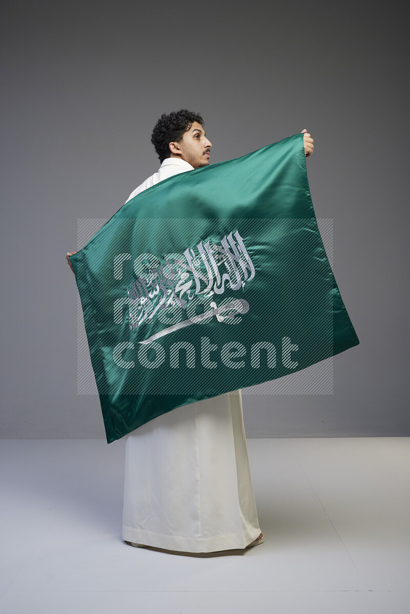 A Saudi man standing wearing thob holding big Saudi flag on gray background