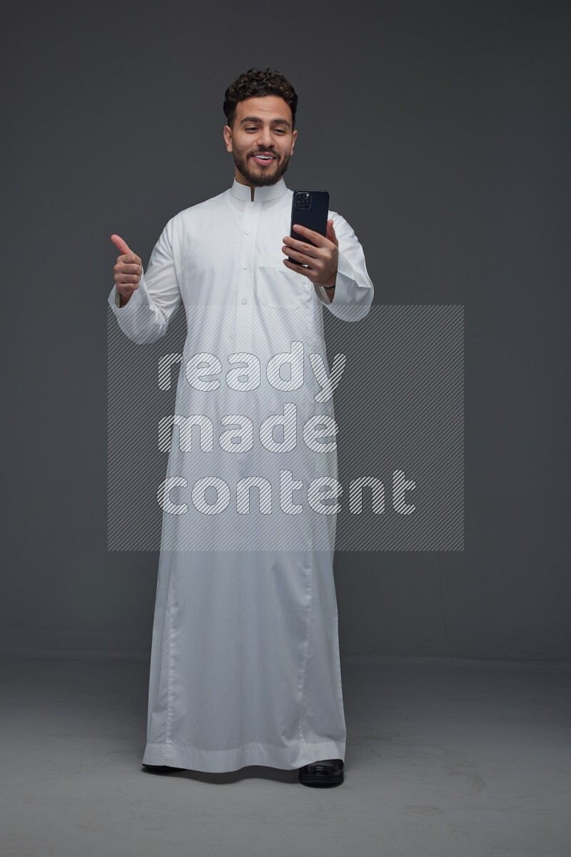 A Saudi man wearing Thobe and making a video call using his phone while standing and making different poses eye level on a gray background