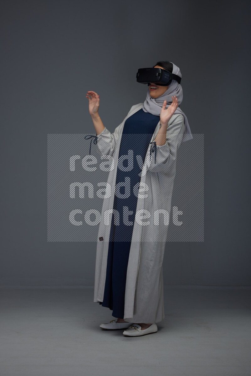 A Saudi woman wearing a light gray Abaya and head scarf standing and wearing VR eye level on a grey background