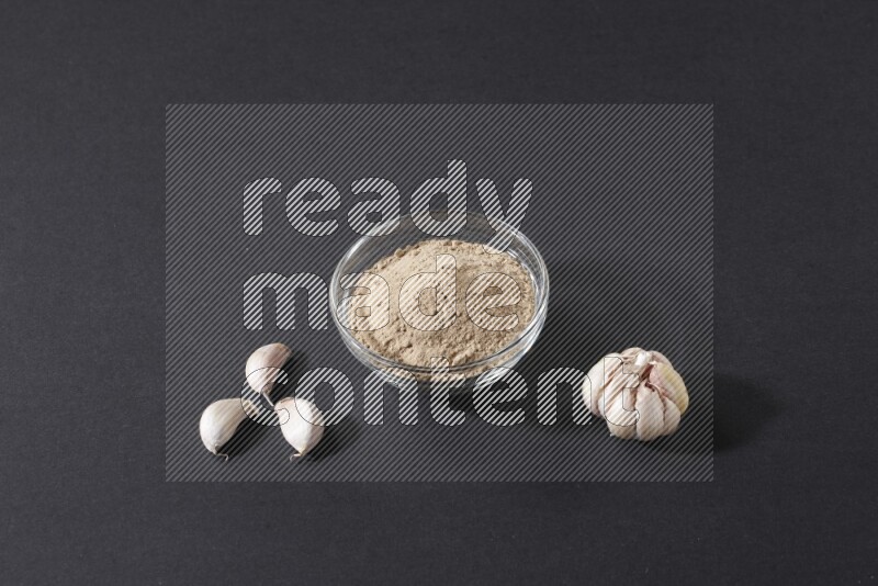 A glass bowl full of garlic powder with garlic bulb and some cloves beside it on a black flooring
