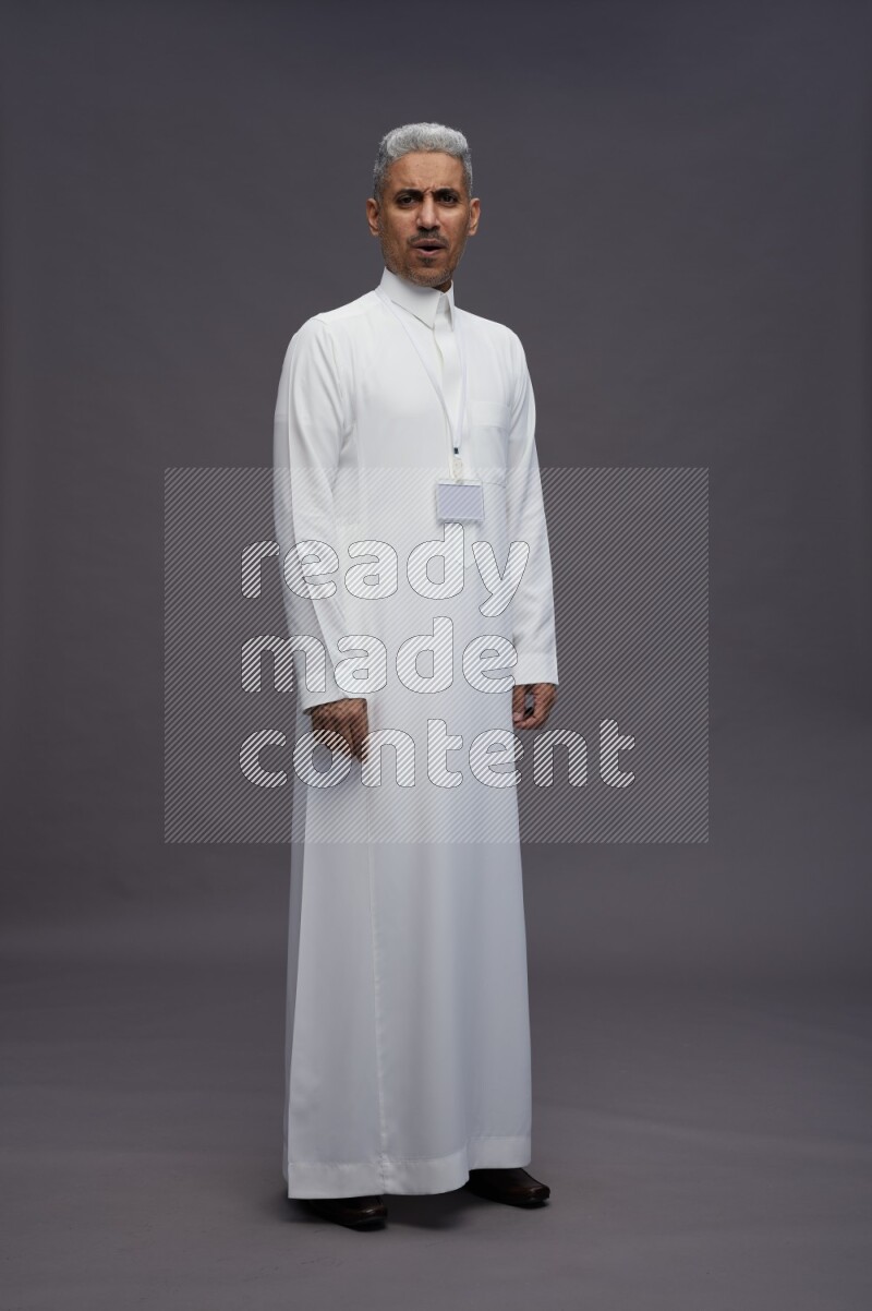 Saudi man wearing thob with neck strap employee badge standing interacting with the camera on gray background