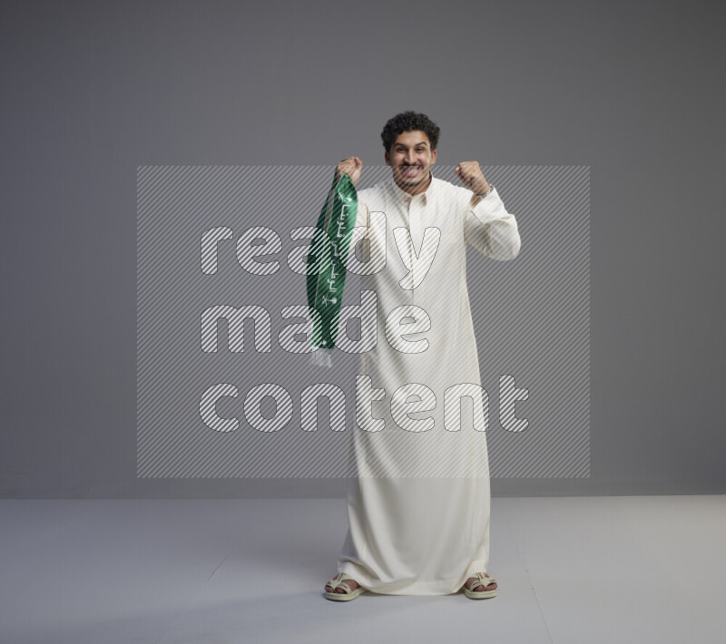 A Saudi man standing wearing thob holding Saudi flag scarf on gray background