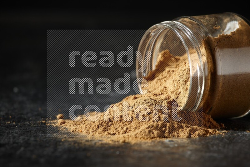 A flipped glass jar full of allspice powder and powder spilled out of it on a textured black flooring